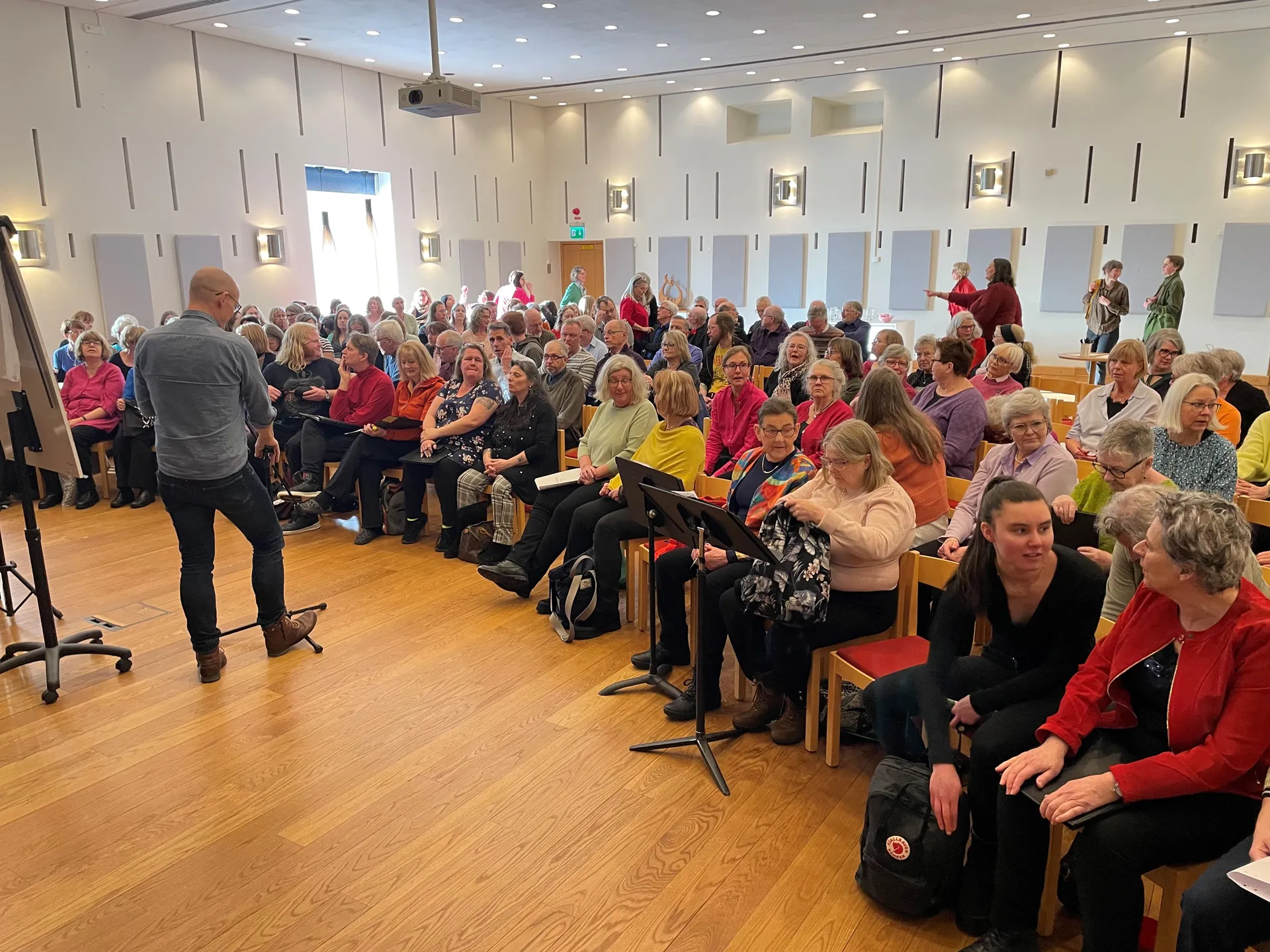 En stor samlingssal med ljusa väggar och trägolv där en person i grå tröja står framför en fullsatt publik sittandes på träbänkar. Salen är upplyst av taklampor och har akustikpaneler på väggarna.