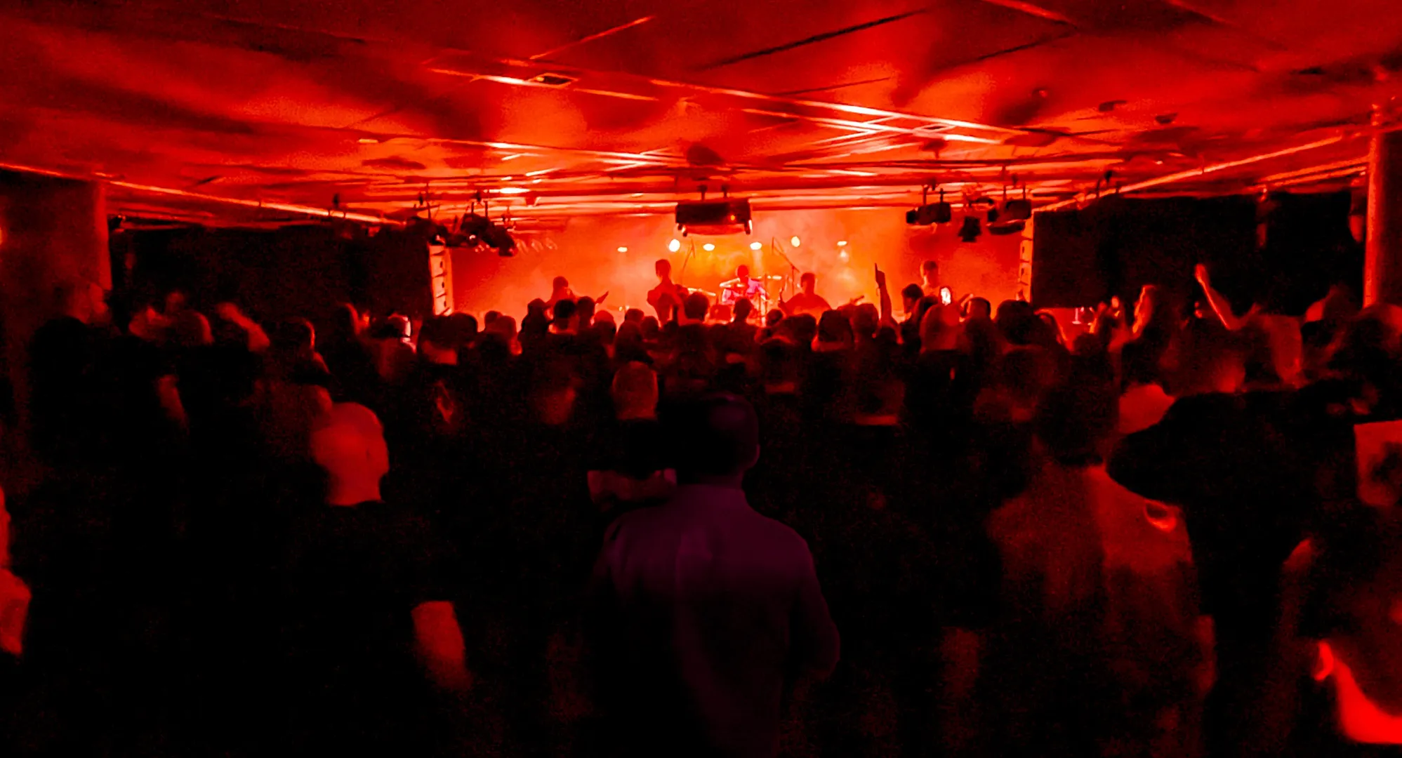 En konsertlokal i mörker med röd belysning där ett band uppträder på scenen framför en stor publik. Taket är täckt med scenbelysning och ljudutrustning.