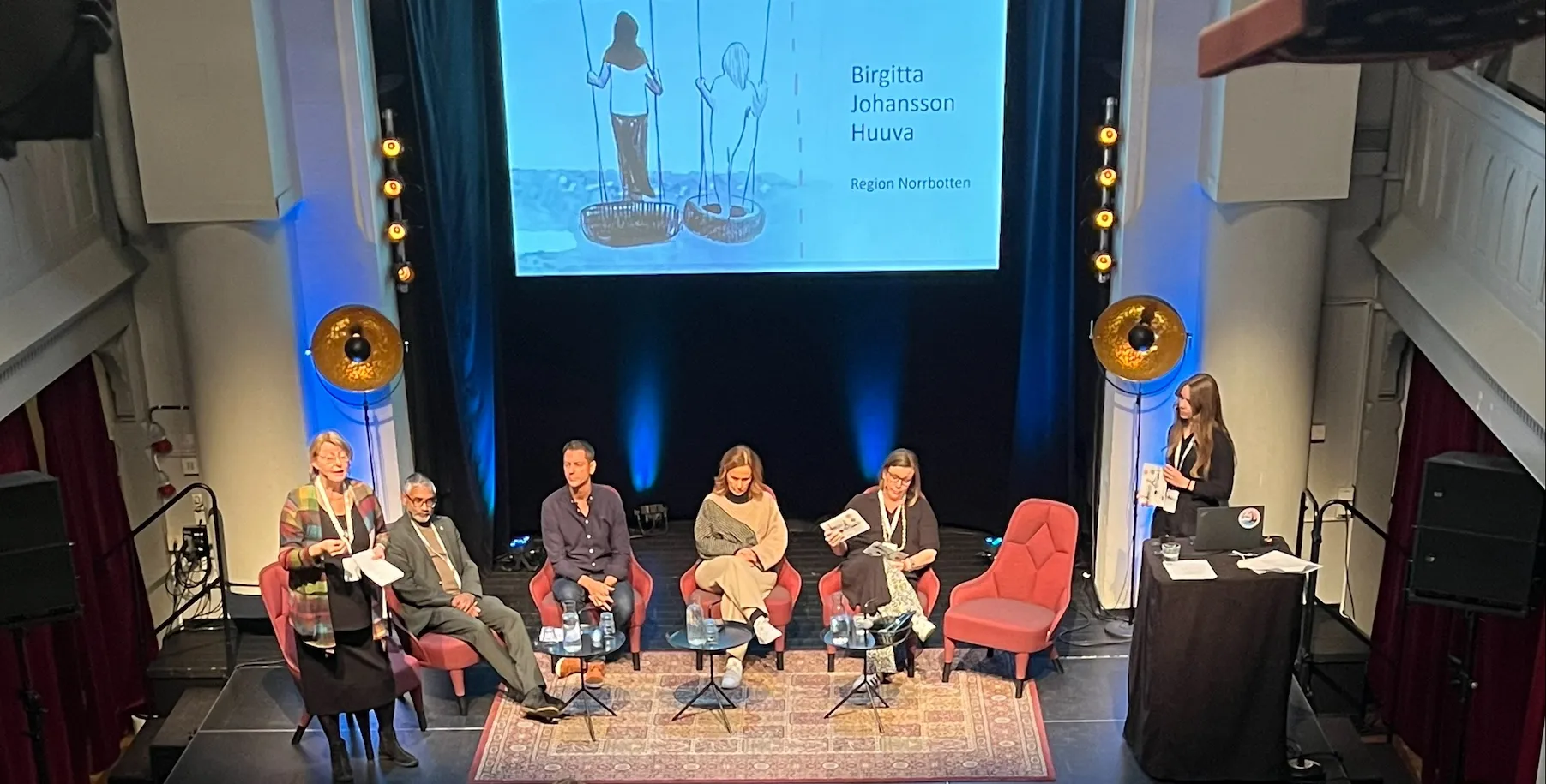 Panel på en scen med sex personer sittande i röda fåtöljer. En presentation visas på storskärm i bakgrunden med namnet Birgitta Johansson Huuva och en illustration av två figurer på gungor. Scenen är dekorerad med blå belysning och orientalisk matta.