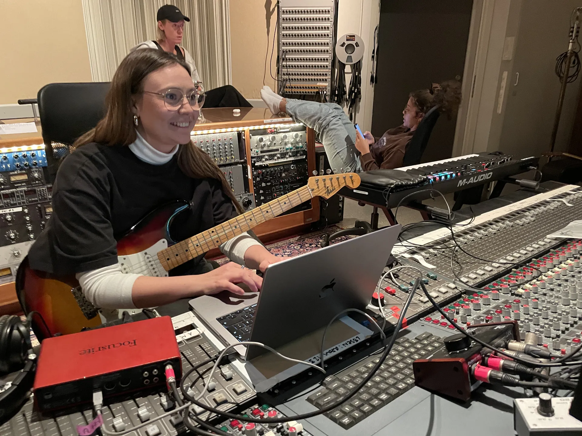 En musikstudio med mixerbord, dator och musikutrustning. En person sitter vid mixerbordet med en elgitarr och arbetar på en bärbar dator. I bakgrunden syns studioutrustning och andra personer.