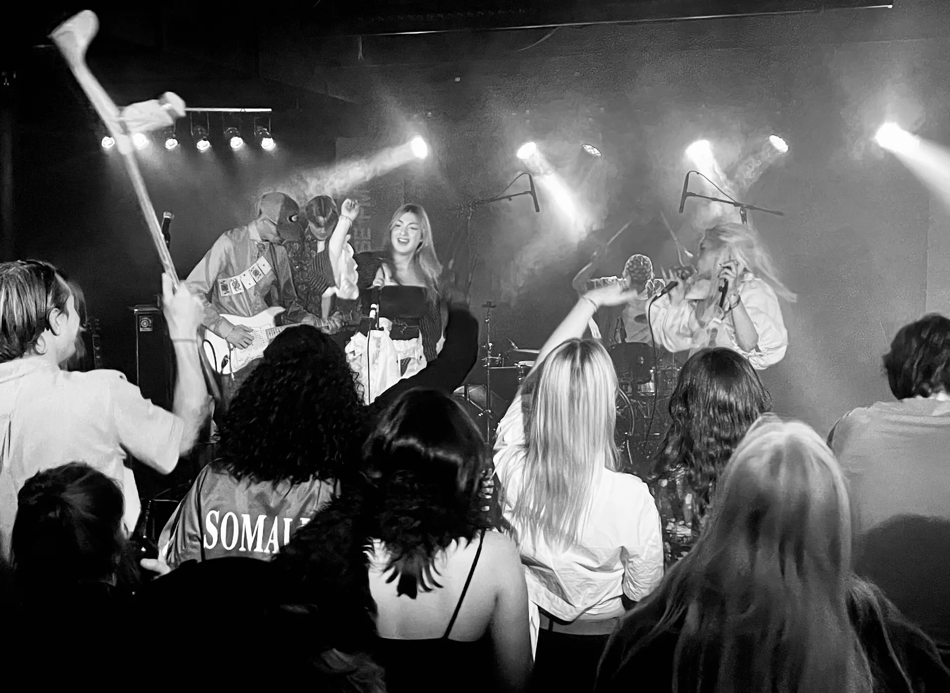 Svartvit konsertbild där ett band uppträder på en rökfylld scen med strålkastare. Publiken syns framifrån med en person som håller upp något i luften. En person i publiken bär en jacka med texten 'SOMA'.