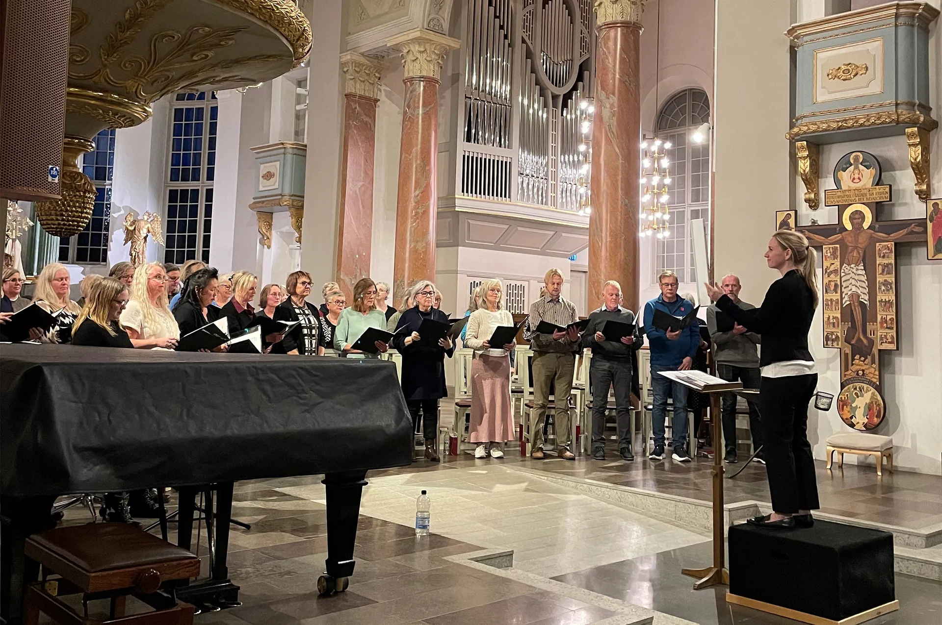 En kör uppställd i en kyrka med en flygel i förgrunden och en dirigent stående på en podium. Kyrkorummet har rosa marmorpelare, en orgel och religiösa konstverk på väggarna.