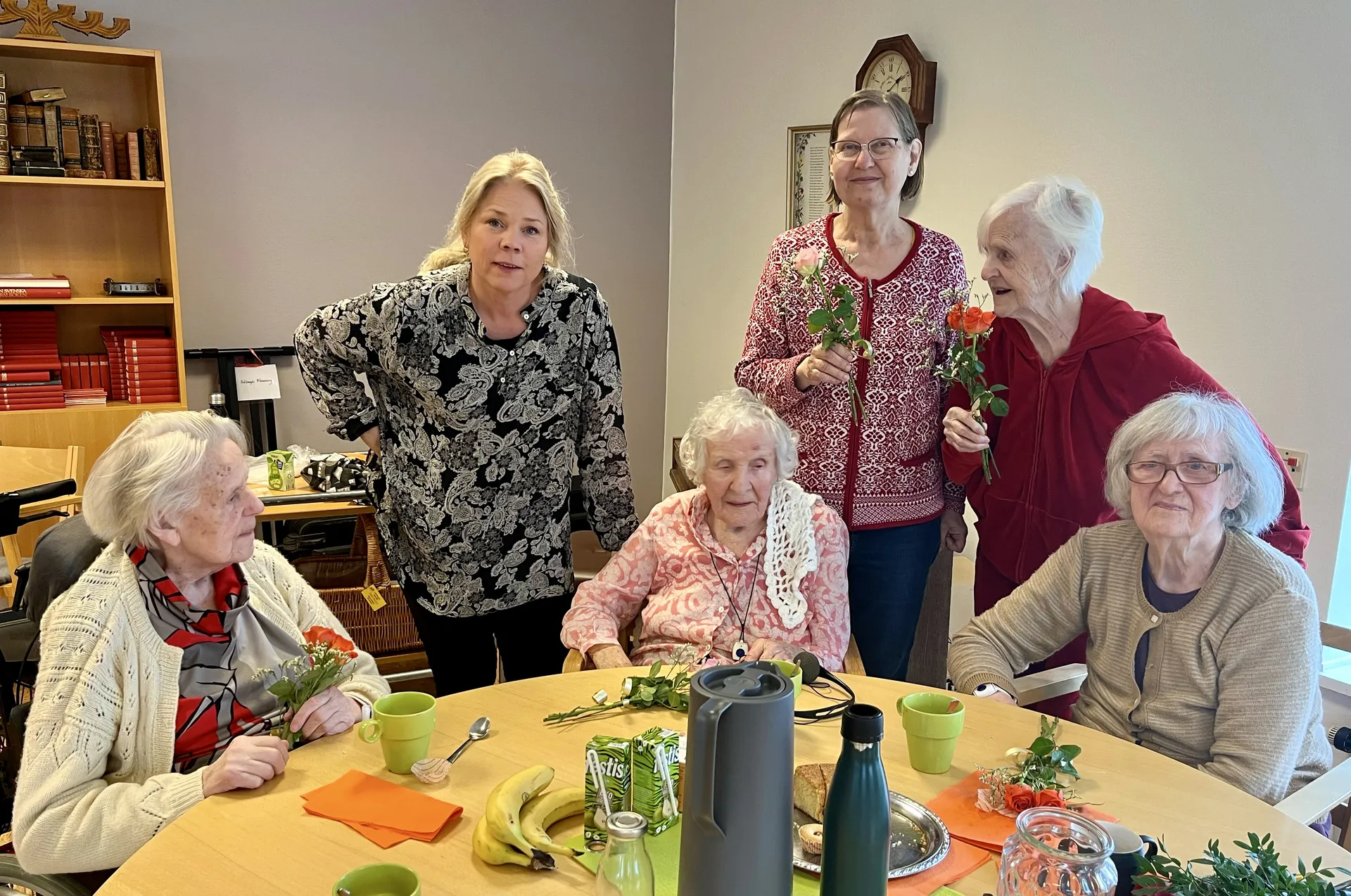 En grupp personer samlade runt ett bord med gula koppar och orange servetter. På bordet finns rosor och förfriskningar. I bakgrunden syns en bokhylla och en väggklocka.
