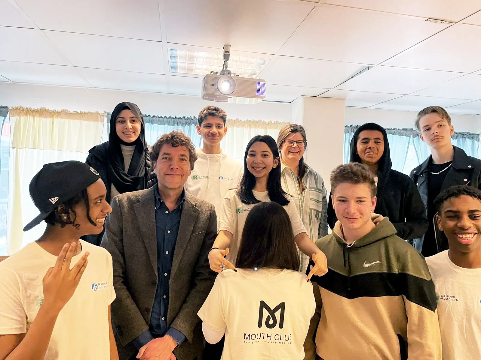 En grupp personer står samlade i ett ljust rum med en projektor i taket. En person i kostym står i mitten medan andra bär t-shirts med 'Mouth Club'-logotypen. Rummet har gardiner och undertaksplattor.