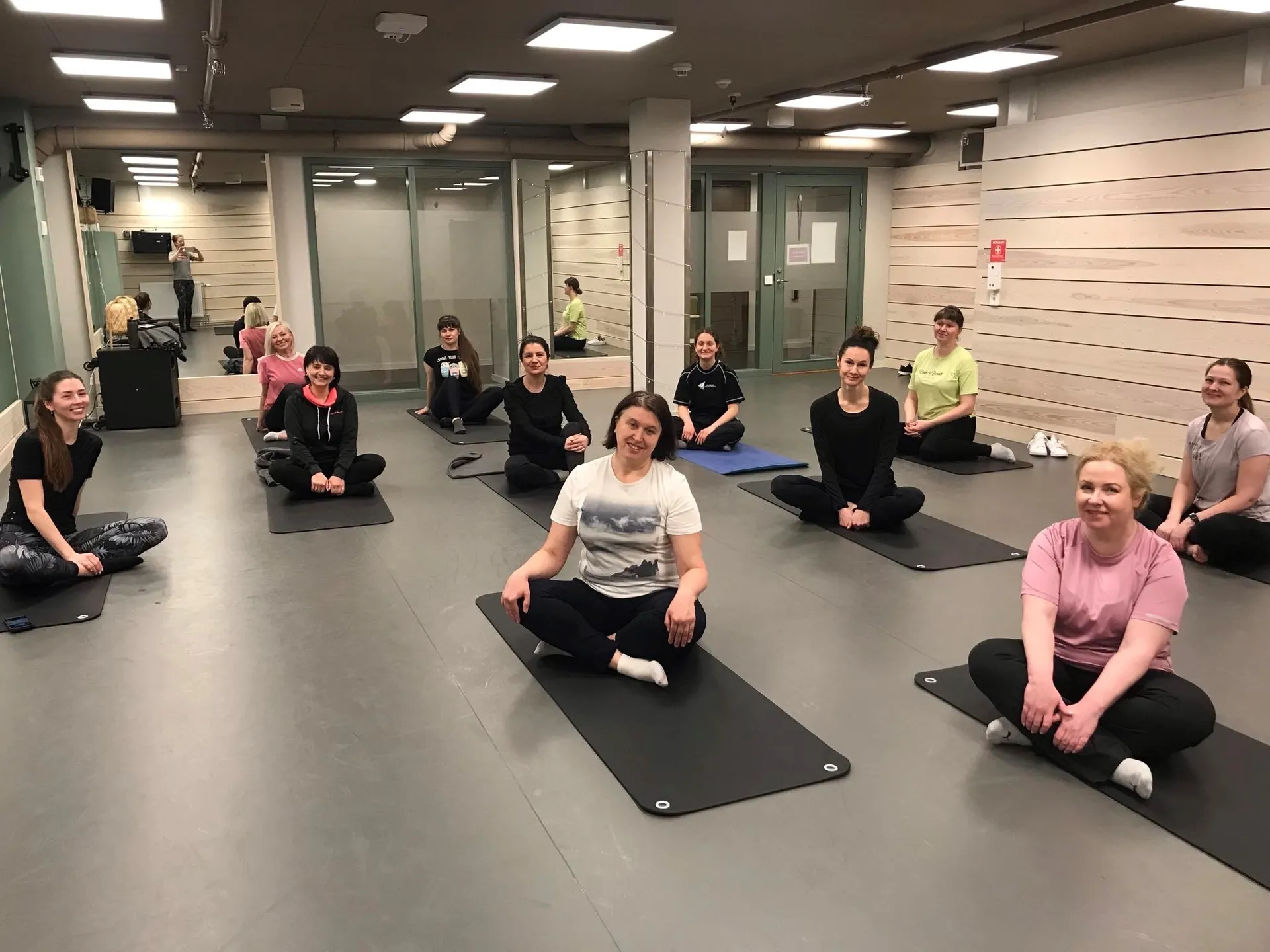 En grupp personer sitter på yogamattor i en träningslokal med ljusa väggar och lysrör i taket. De är placerade med avstånd från varandra i ett mönster över golvet.