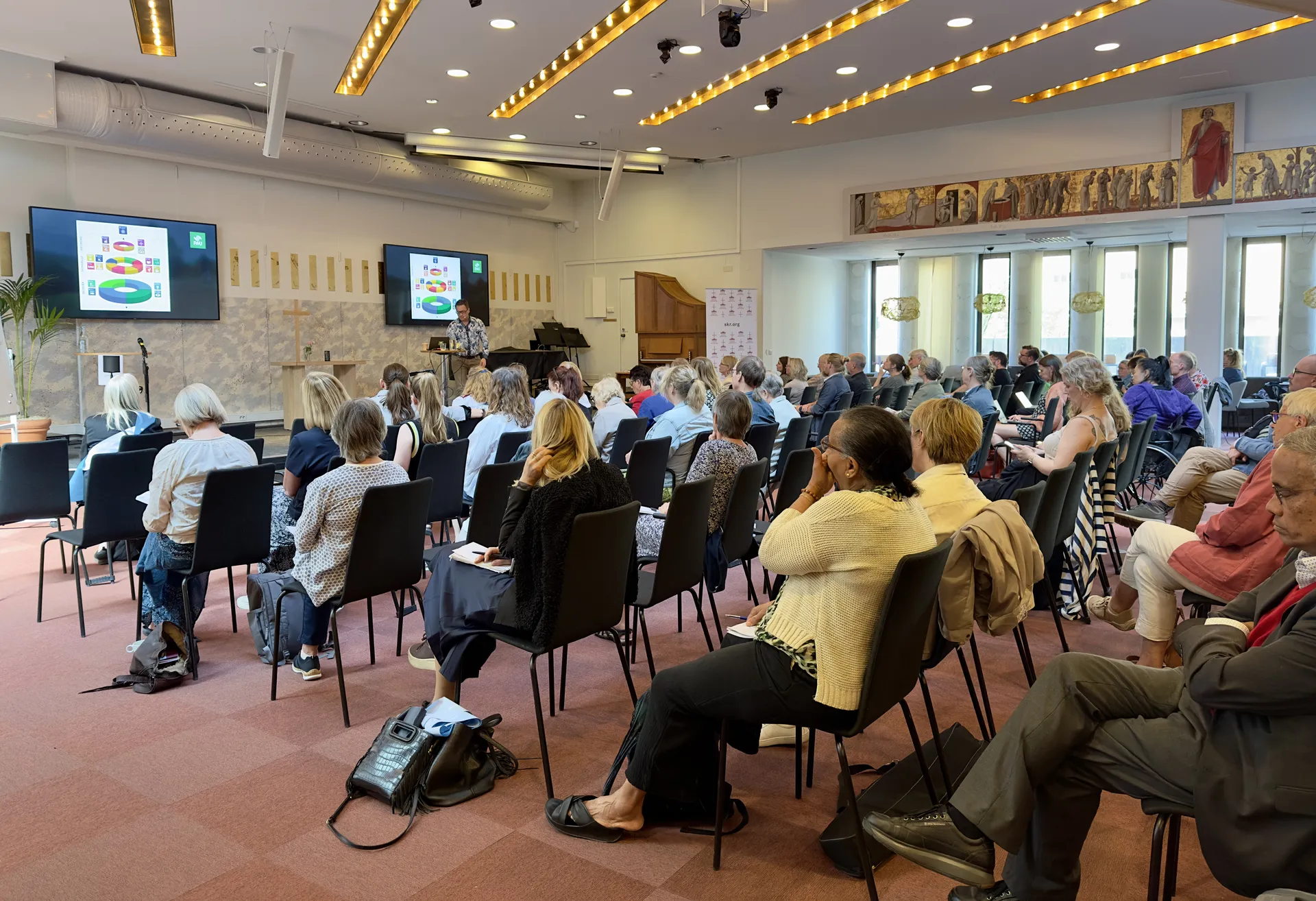 Konferensrum med publik som sitter på svarta stolar och tittar på en presentation som visas på två stora TV-skärmar. Rummet har guldaktiga ljusskenor i taket och religiösa konstverk på väggen.