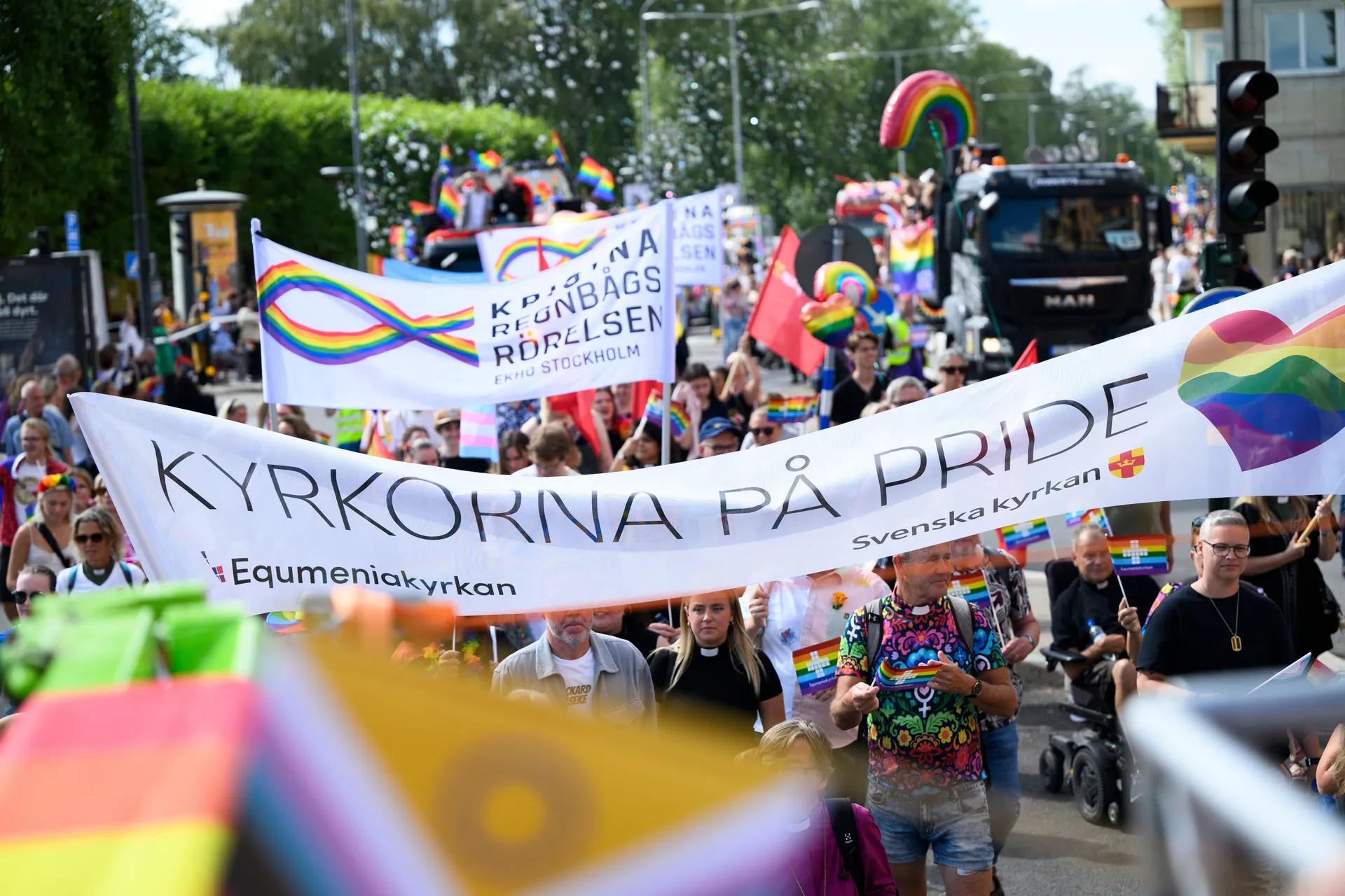 Pride-parad med banderoller från Svenska kyrkan och andra religiösa organisationer. Många regnbågsflaggor och människor som deltar i paraden syns i bilden.