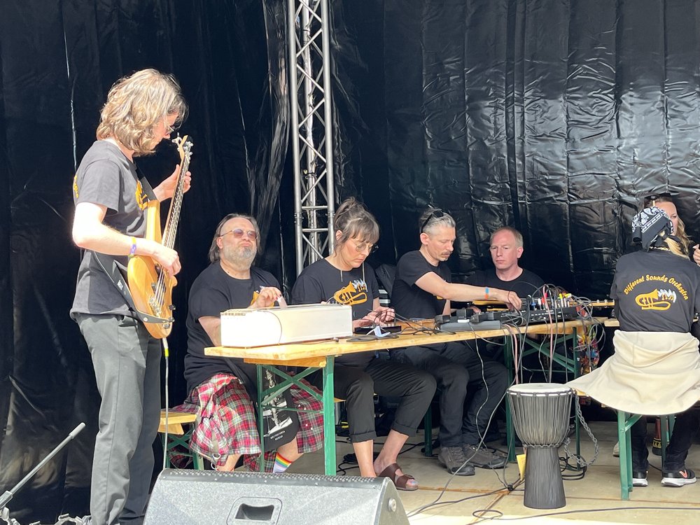 Deltagare från Medis 5 på en scen en solig dag i Polen. En deltagare spelar bas medan de andra sitter bakom ett mixerbord.