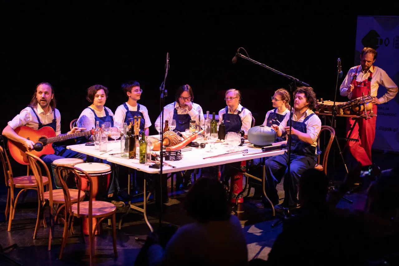 En grupp musiker sitter vid ett långt bord på en mörk scen, alla klädda i blå förkläden och vita skjortor. En person spelar gitarr och det finns flera flaskor och glas på bordet. Scenen är upplyst med teatraliskt ljus.