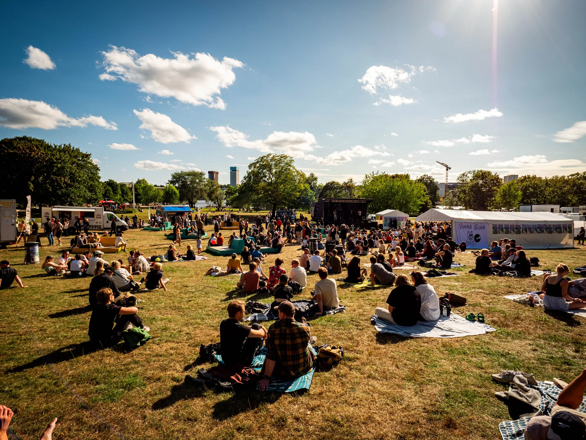 En solig sommardag på en utomhusfestival där många människor sitter på filtar i gräset framför en scen. I bakgrunden syns food trucks, tält och höga byggnader mot en klarblå himmel med vita moln.