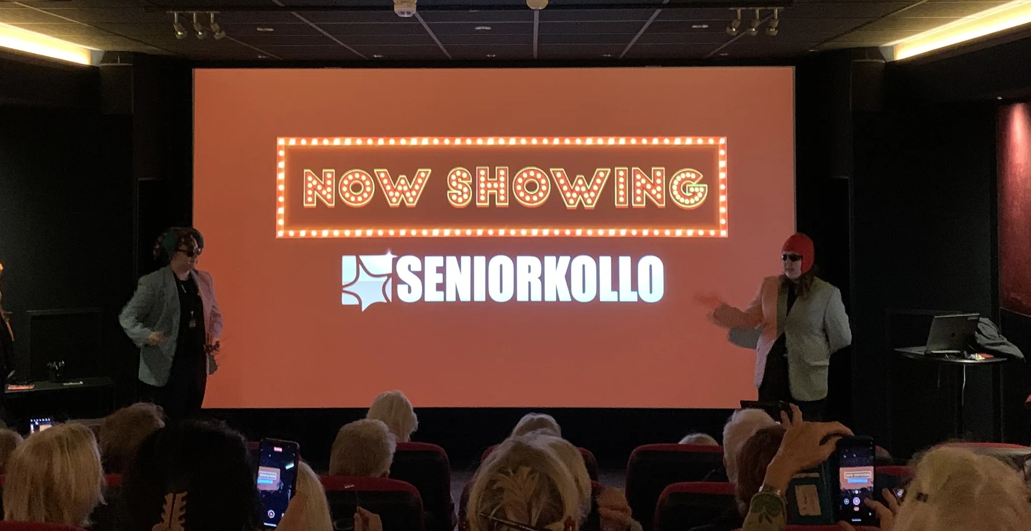Biografsalong med publik där två personer står framför en stor projektorduk som visar texten 'NOW SHOWING' i upplysta bokstäver och 'SENIORKOLLO' i vitt