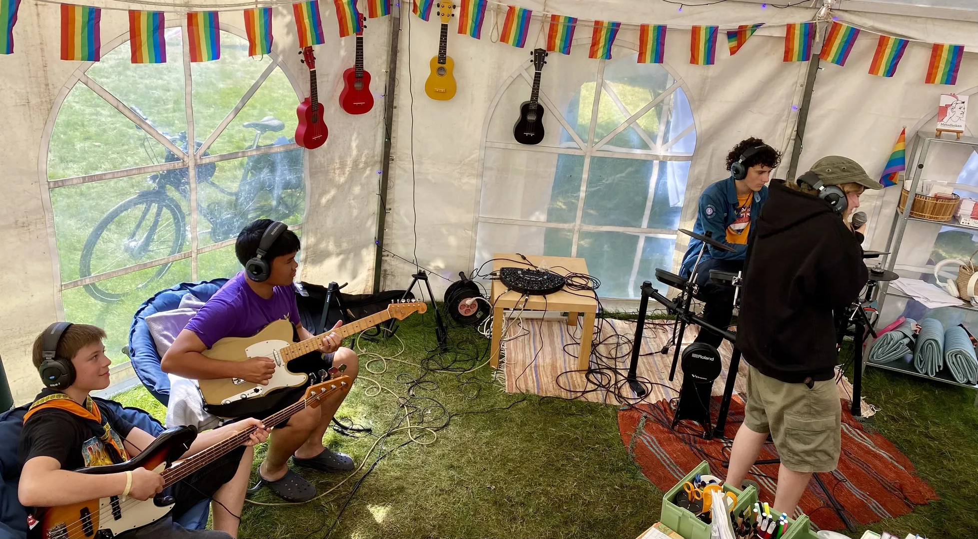 Musiker spelar instrument i ett vitt partytält dekorerat med prideflaggor. Ukuleles hänger på väggen och musikutrustning är utspridd på golvet.