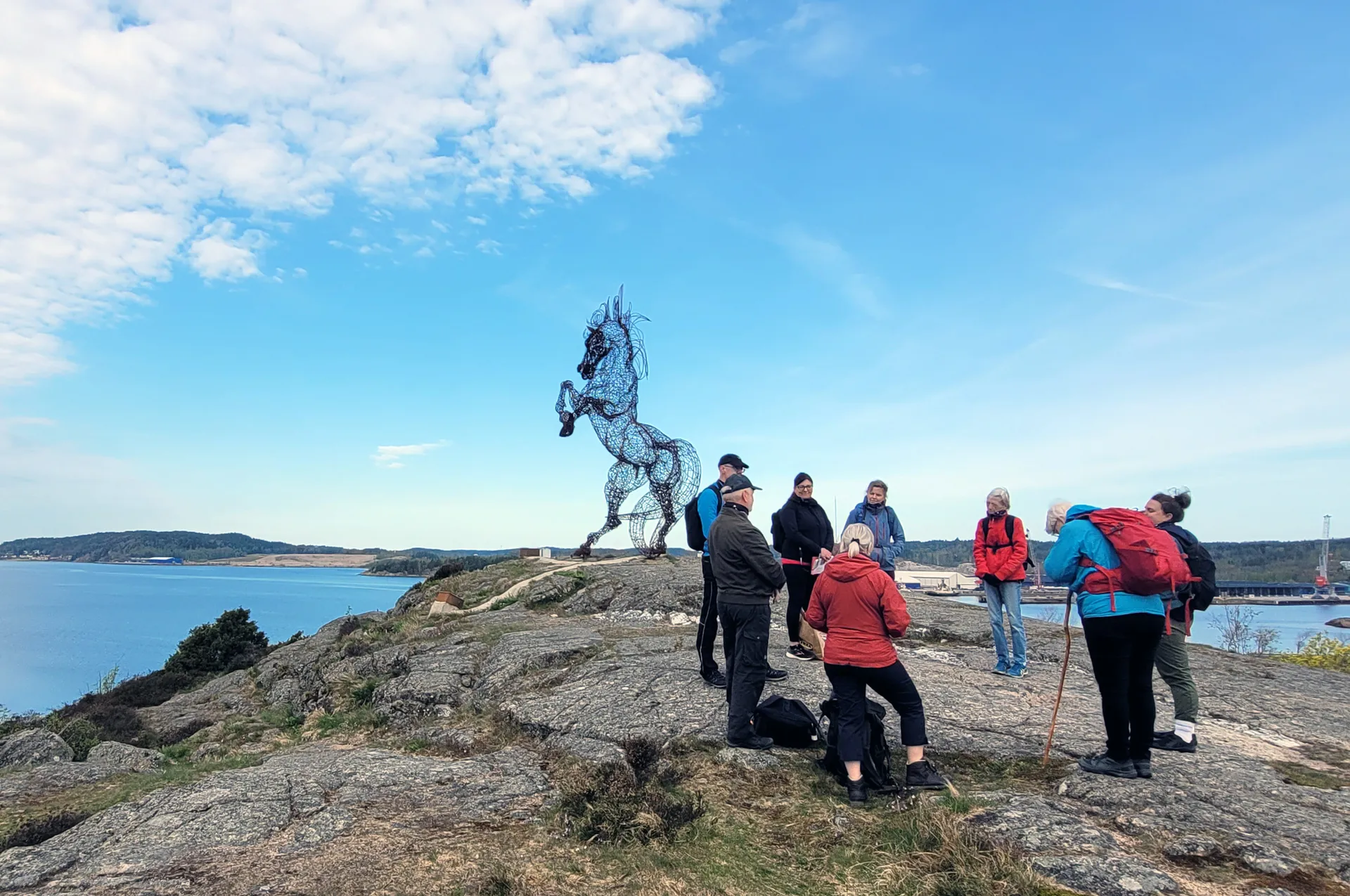 En grupp vandrare står på en klippa vid havet och betraktar en stor trådkonstinstallation i form av en stegrande häst. I bakgrunden syns hav, öar och en blå himmel med vita moln.