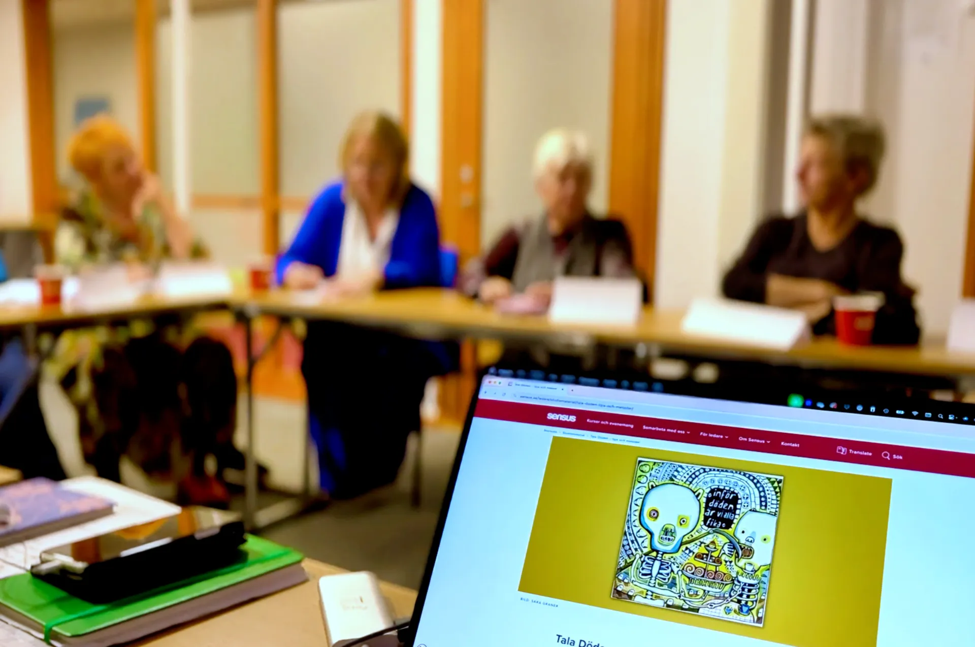 Ett mötesrum där personer sitter runt ett bord med en bärbar dator i förgrunden som visar en webbsida med en tecknad skelettfigur på gul bakgrund