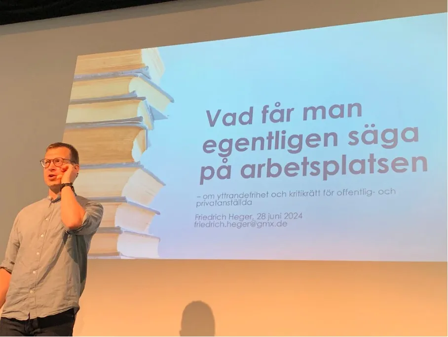 En presentationsbild med texten 'Vad får man egentligen säga på arbetsplatsen' mot en ljusblå bakgrund. Till vänster syns en stapel med böcker. En person står framför presentationen i en grå skjorta.