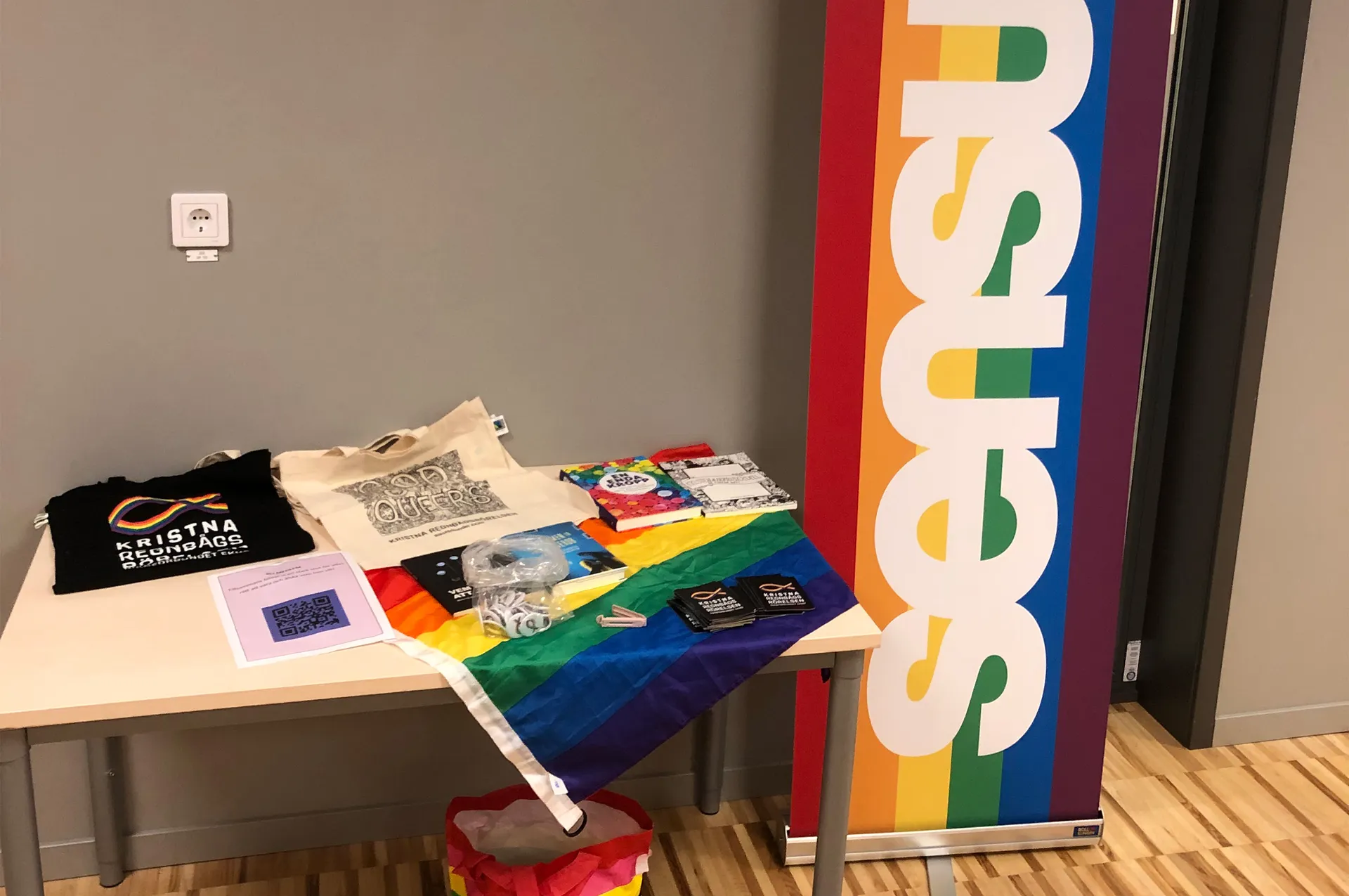 Ett bord med PRIDE-relaterade föremål inklusive en regnbågsflagga, tygkassar och diverse merchandise. Bredvid bordet står en färgglad roll-up banner med regnbågsfärger.