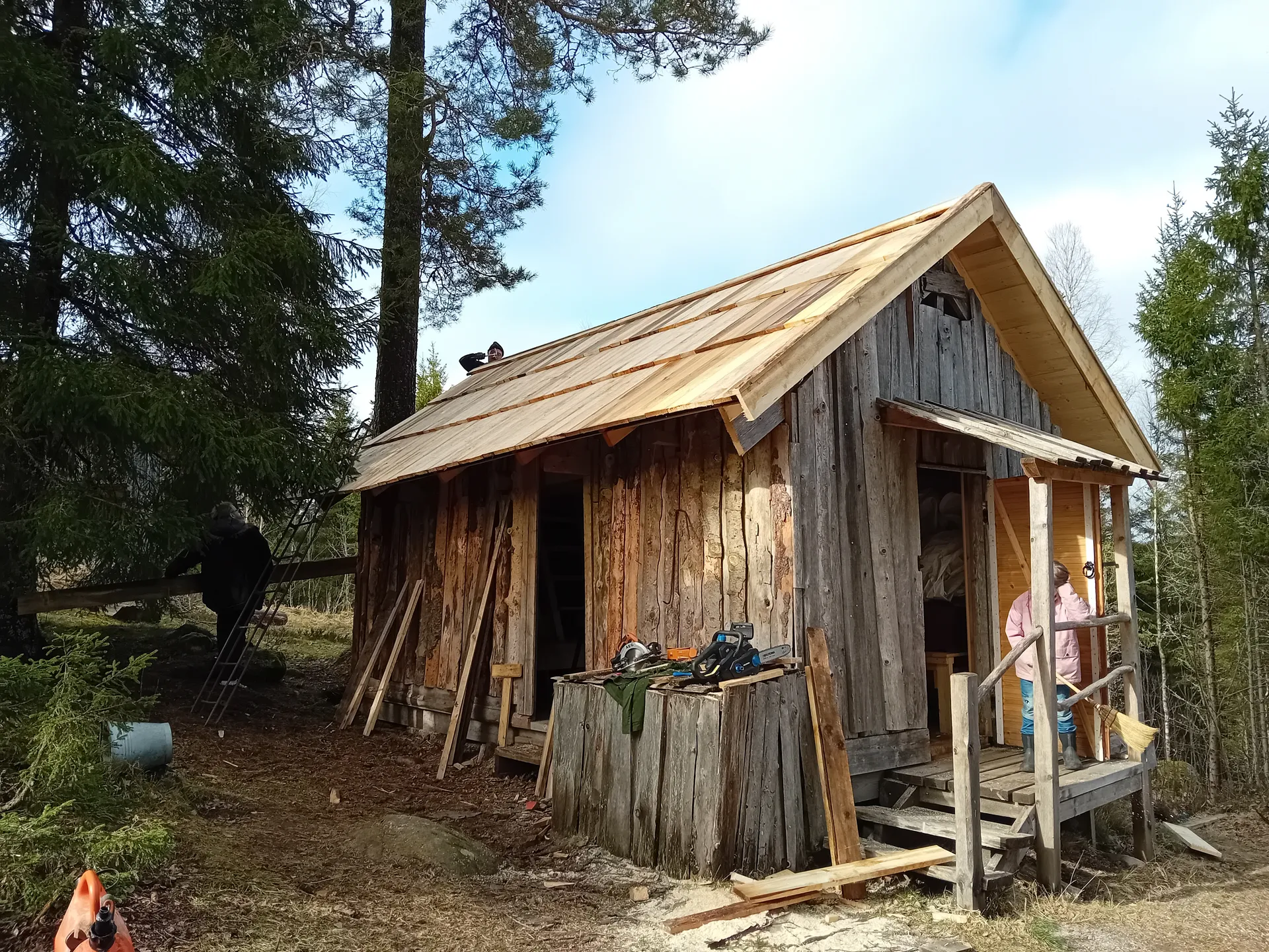 En gammal timmerstuga med nytt trätak under renovering. På verandan ligger byggverktyg och material. Stugan är omgiven av barrträd i en skogsglänta.