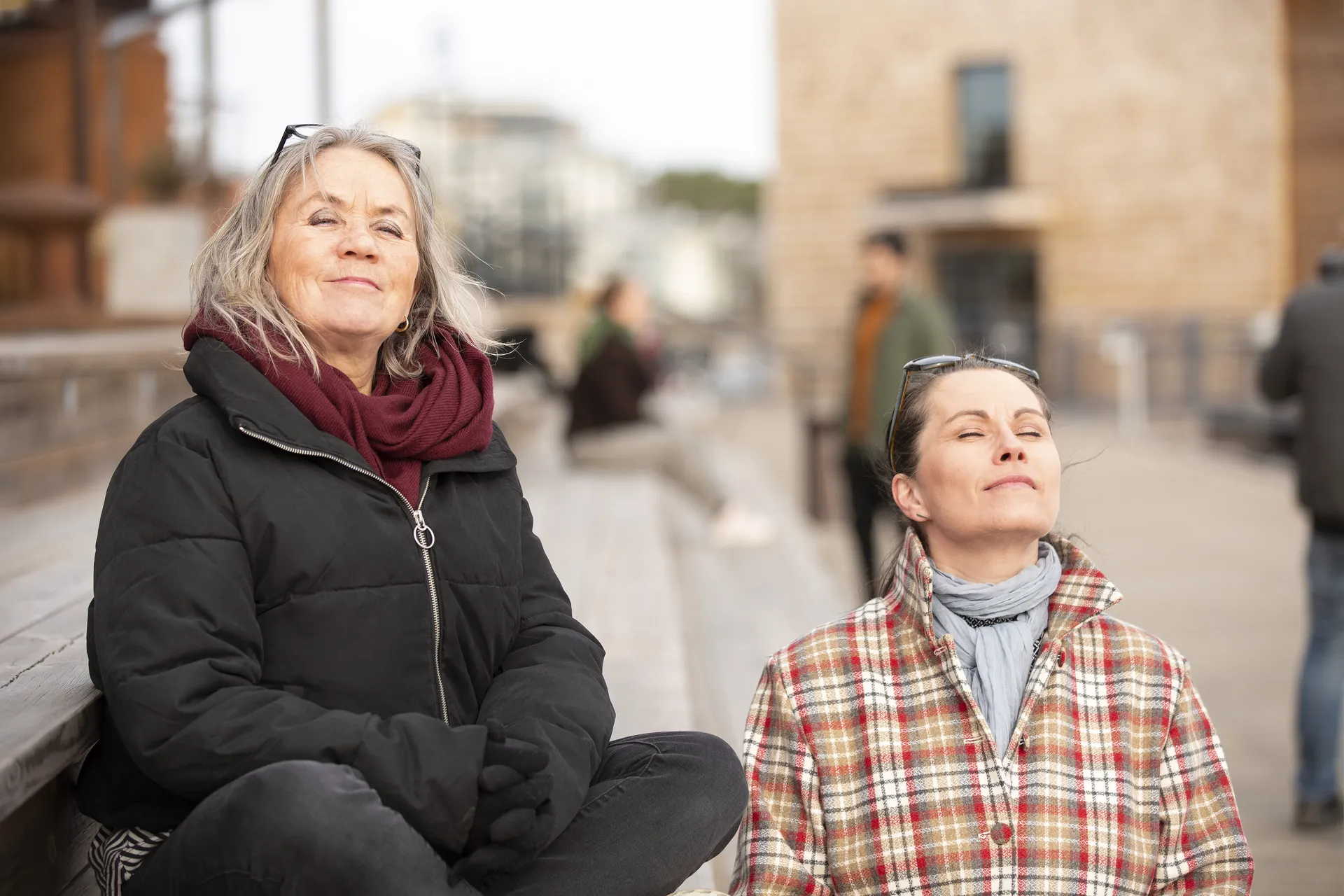 Två personer sitter utomhus och njuter av solen med slutna ögon. En har på sig en svart vinterjacka med vinröd halsduk, den andra bär en rutig flanellskjorta med grå halsduk. I bakgrunden syns en stadsmiljö med beigea byggnader.