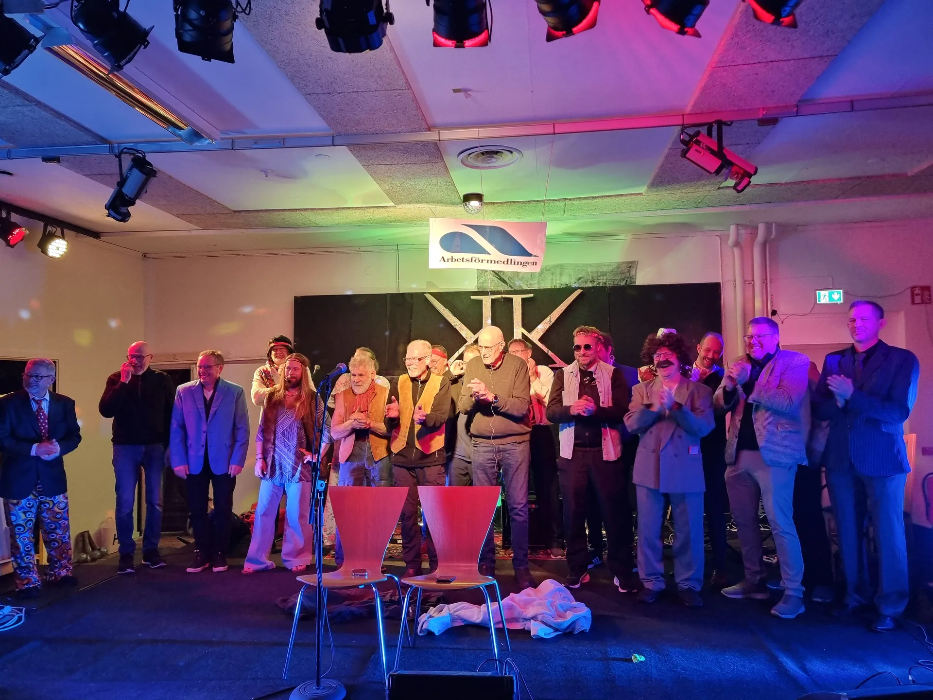 En grupp personer står på en scen med färgad belysning i rosa, blått och grönt. På väggen syns en Arbetsförmedlingen-logotyp. Två röda stolar står framför scenen och i taket hänger teaterlampor.