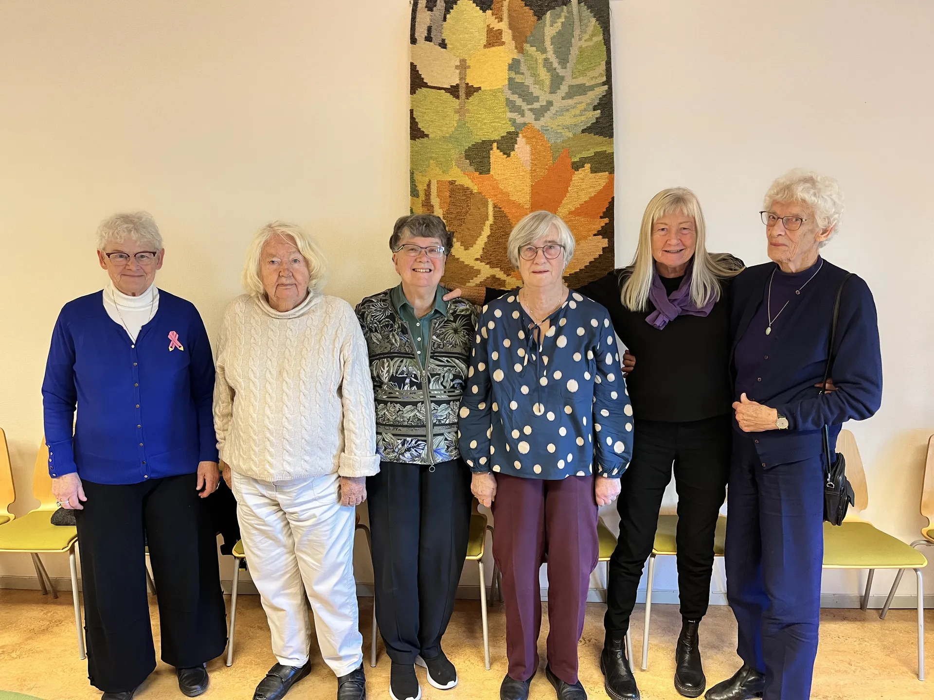 Sex äldre personer står på rad framför en vägg med en stor höstinspirerad väggbonad. De bär olika färgglada kläder och står framför gula stolar. På väggen bakom dem syns en stor textilkonst med löv i höstfärger.