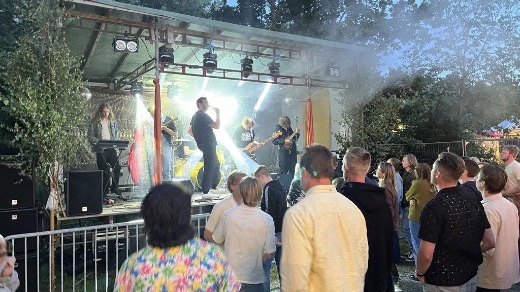 Ett band uppträder på en utomhusscen med strålkastare och scenbelysning. Publiken står framför scenen bakom ett staket. Scenen är utrustad med högtalare och ljusrigg.