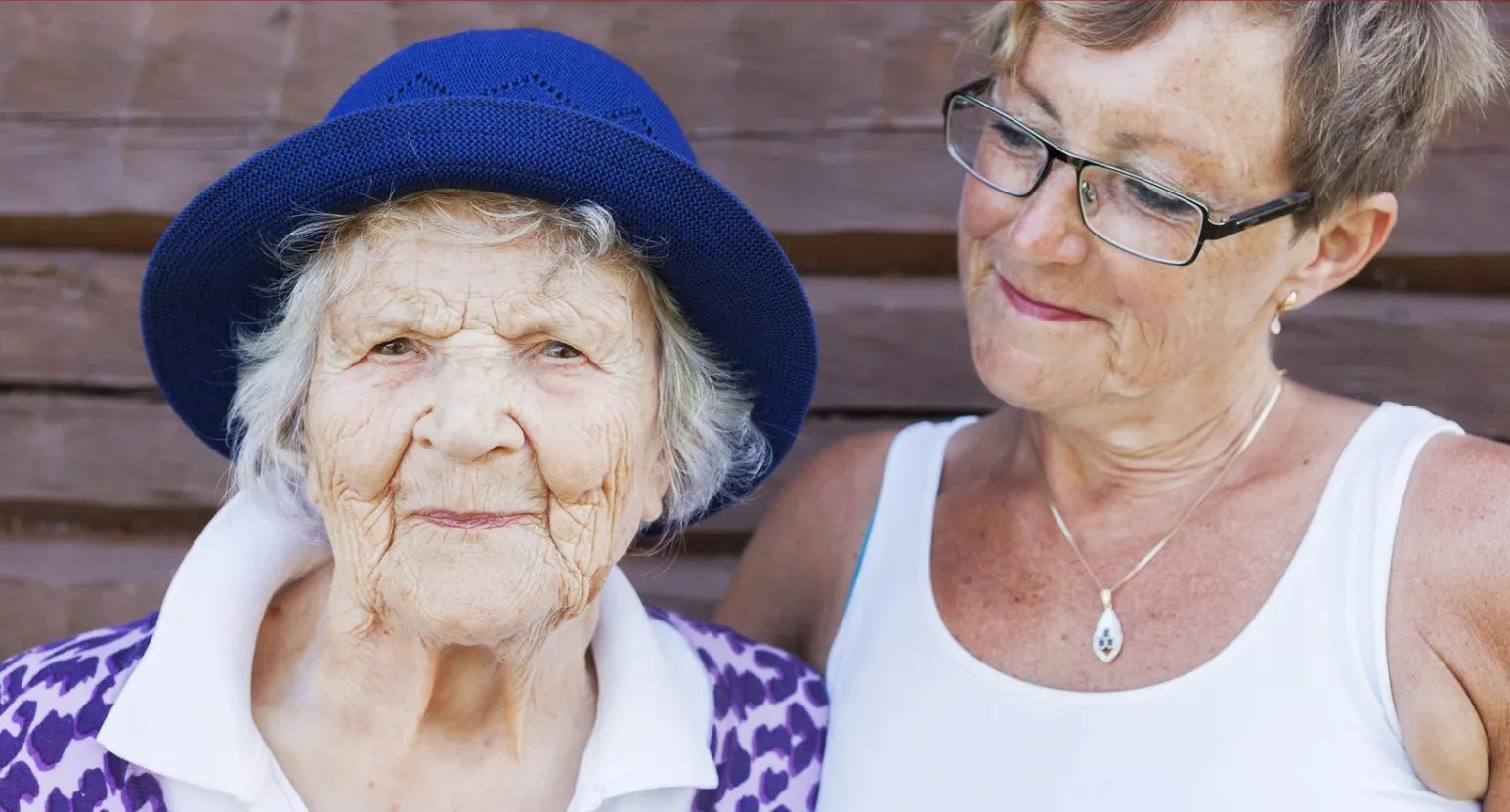 Två personer sitter tillsammans utomhus mot en trävägg. En äldre person bär en blå hatt och en lila-vit mönstrad tröja, medan den andra har på sig ett vitt linne och glasögon.