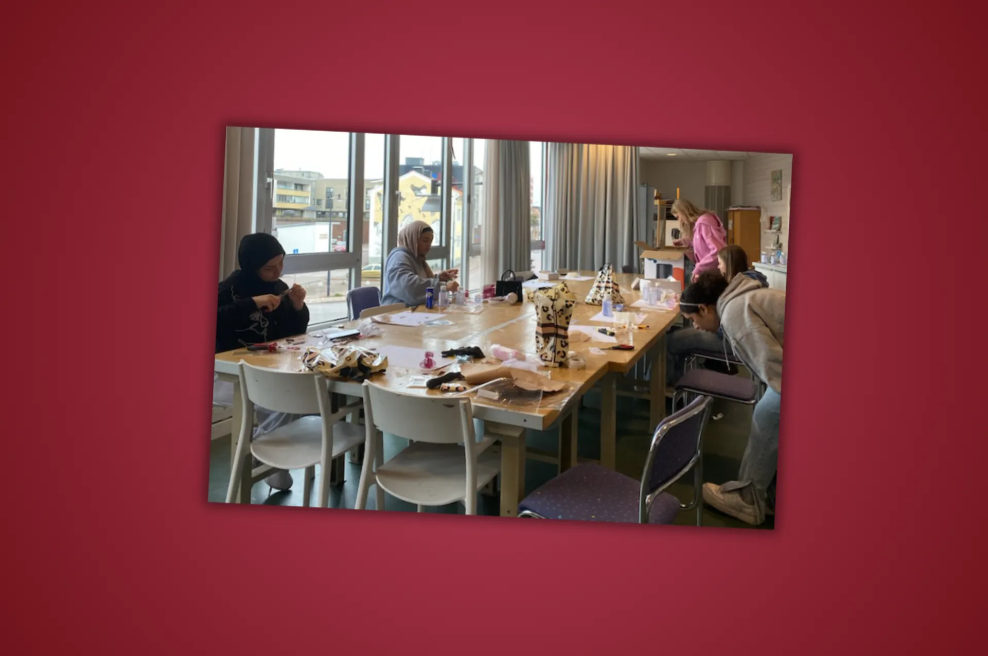 Personer sitter runt ett långt bord i ett ljust rum med stora fönster och arbetar med någon form av hantverk eller pyssel. På bordet ligger diverse material utspridda.