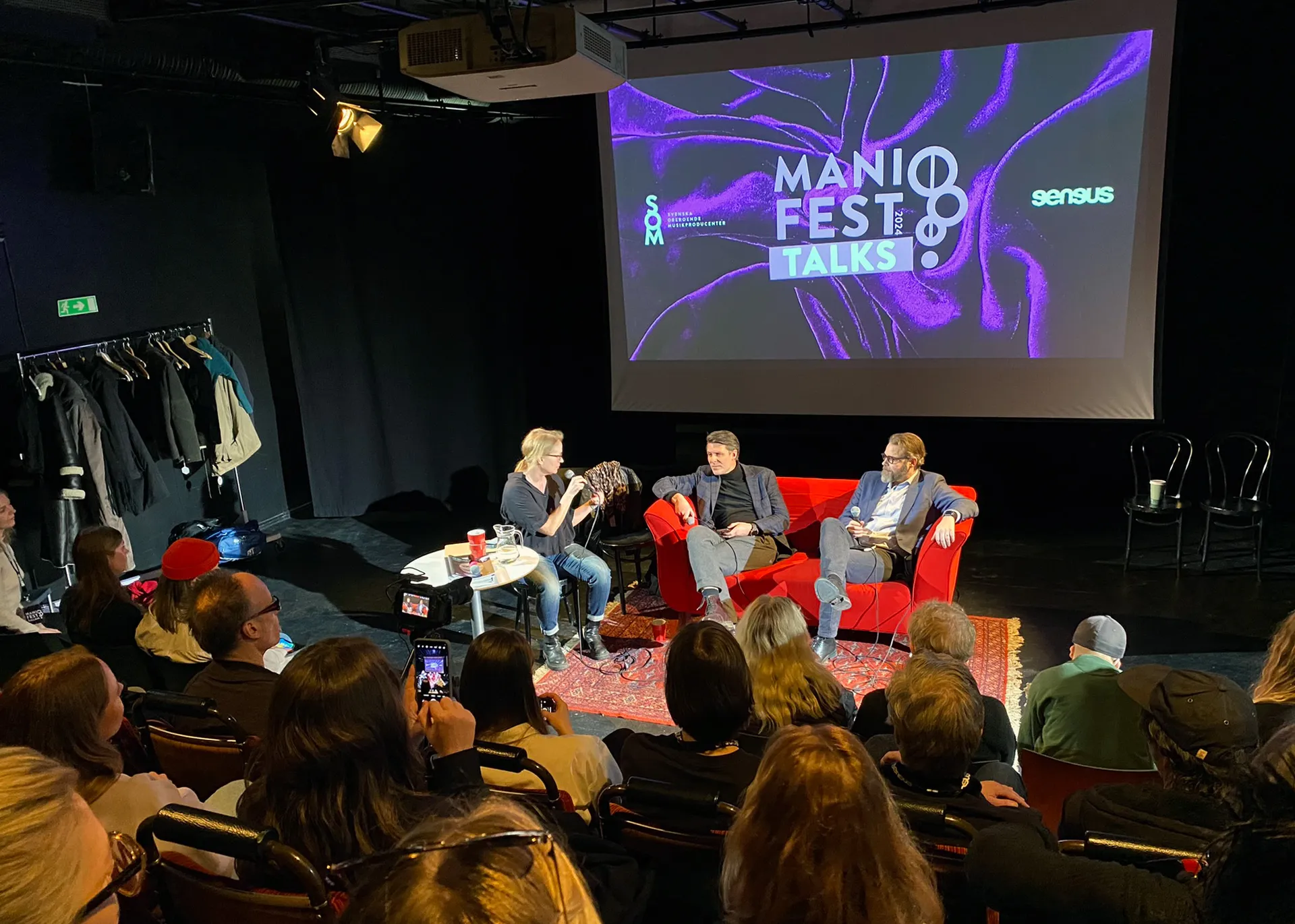 Scen från ManiFest Talks med tre personer som sitter i röda fåtöljer på en upplyst scen framför en stor projektorduk som visar festivalens logotyp i lila och vitt. Publiken sitter i mörka teaterstolar framför scenen.