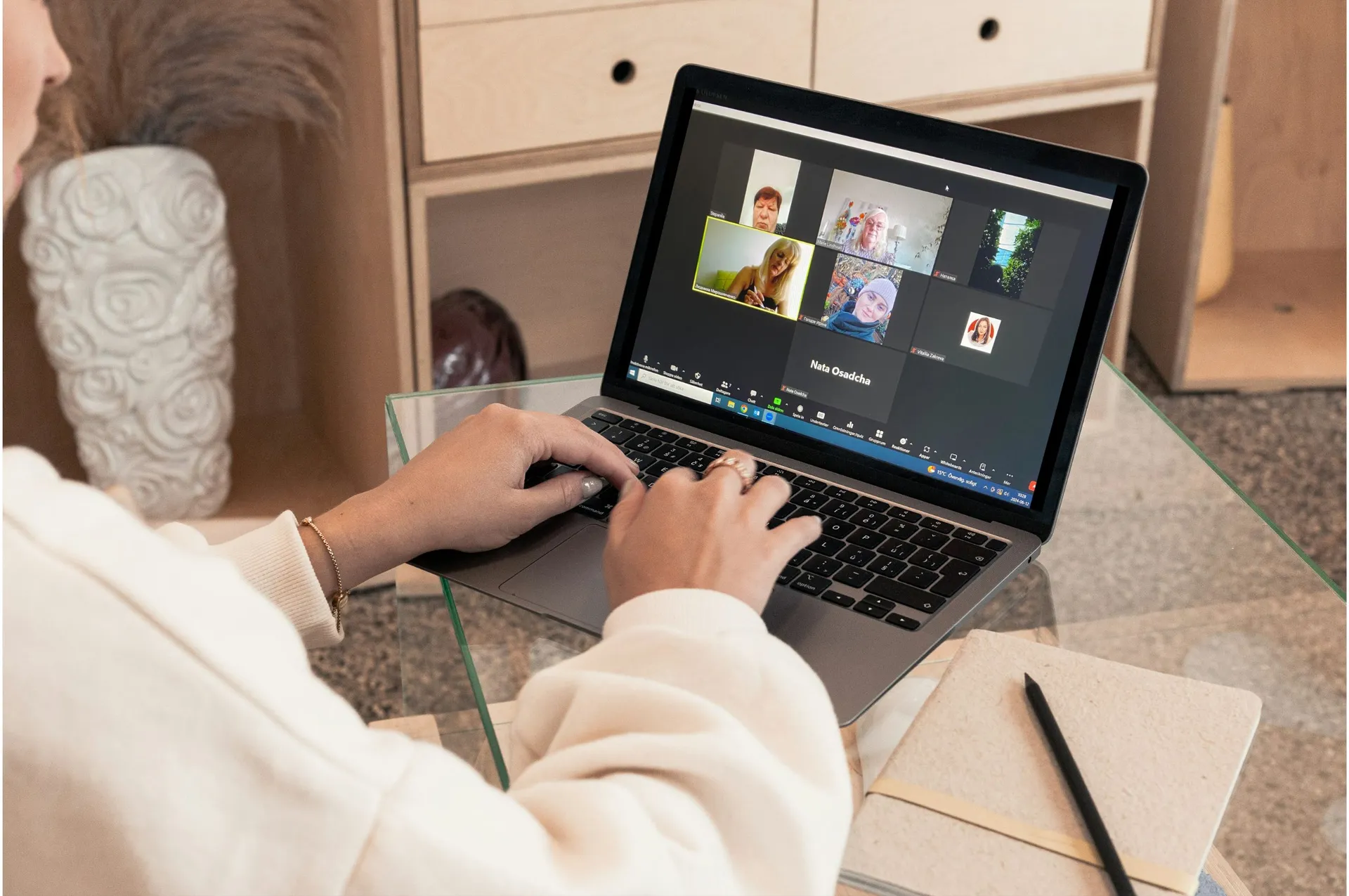 Person i vit tröja sitter vid ett glasbord och använder en MacBook för ett videosamtal. På skärmen syns flera deltagare i ett Zoom-möte. Bredvid datorn ligger ett anteckningsblock med en penna.